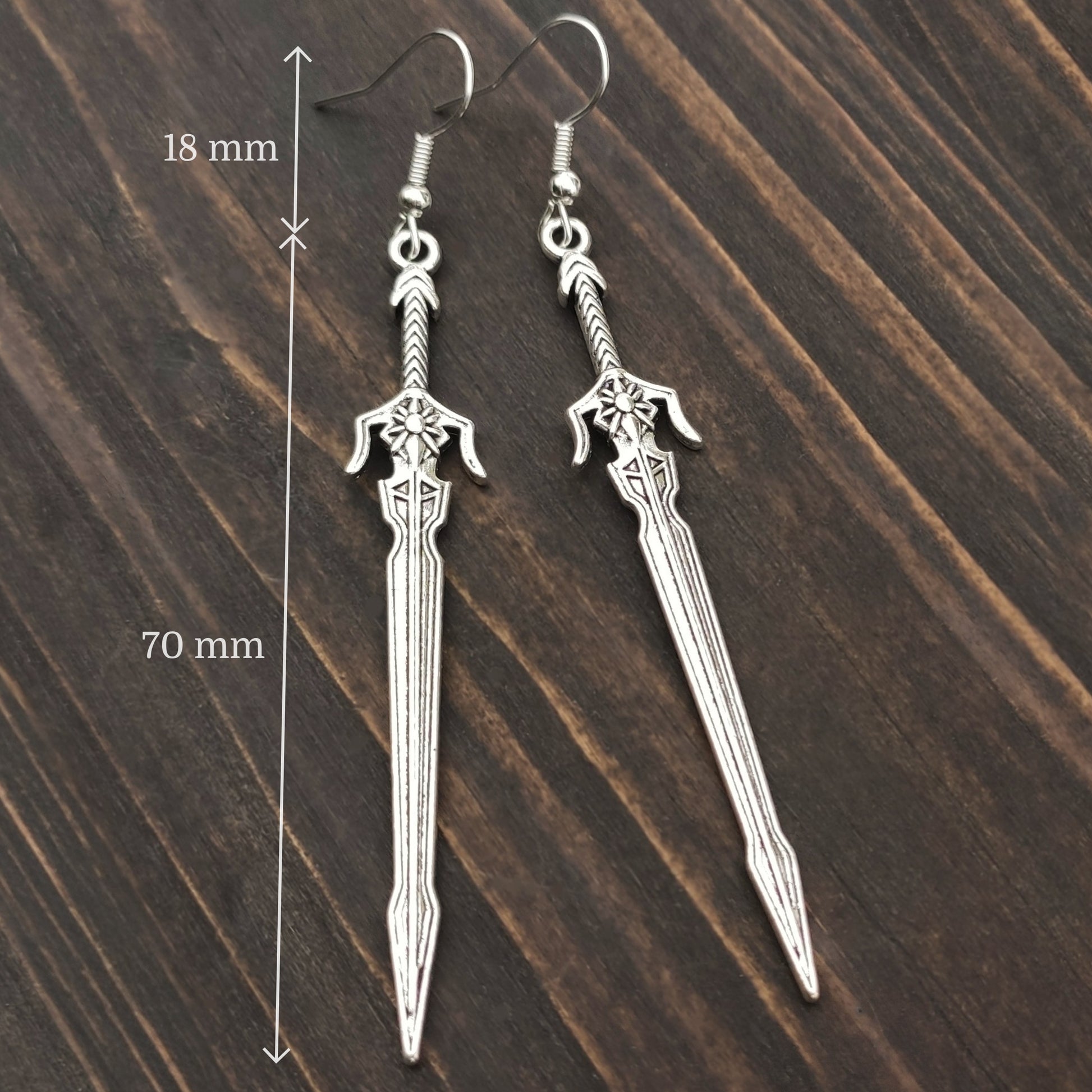 Silver sword earrings with hooks on a wooden surface with measurements indicated.