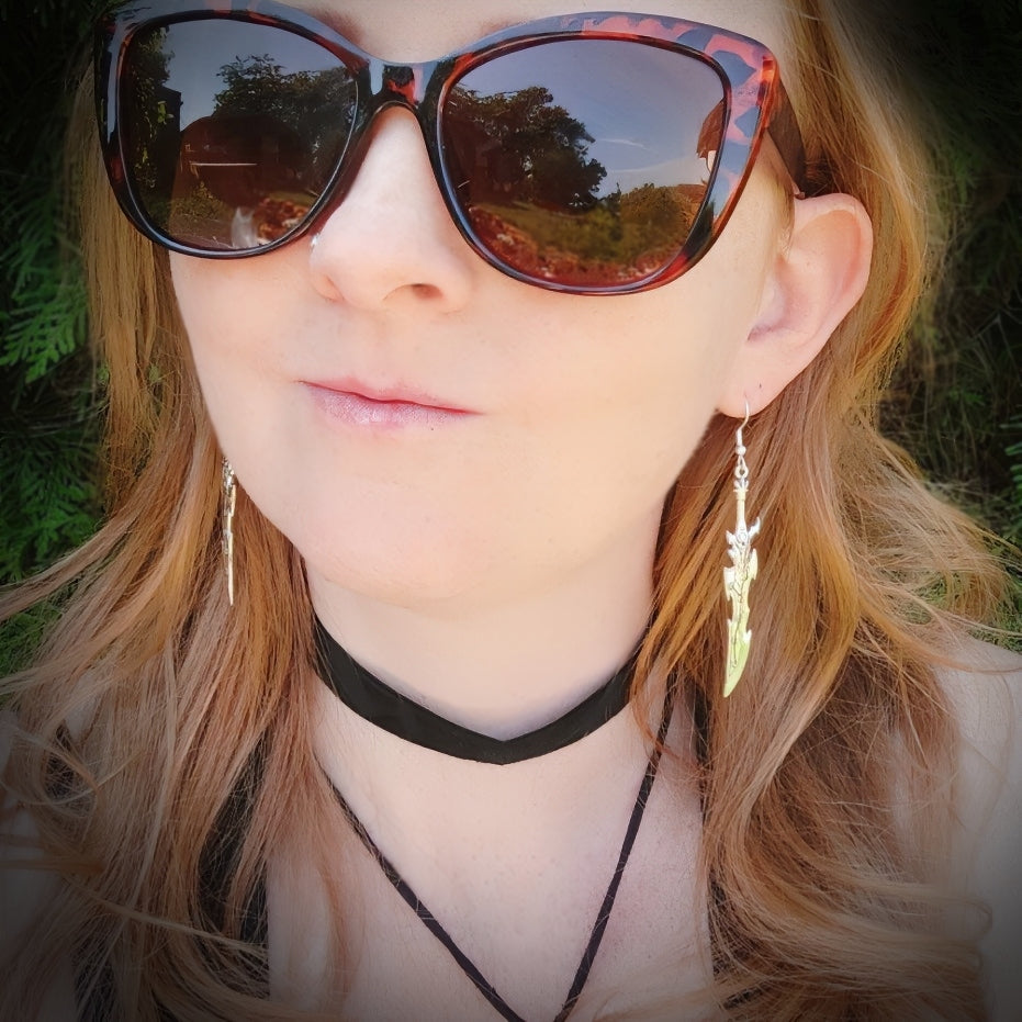 A female model wearing long sword earrings with a curved and spiky blade design. The silver swords look like they belong in a fantasy space battle and have stainless steel hooks.