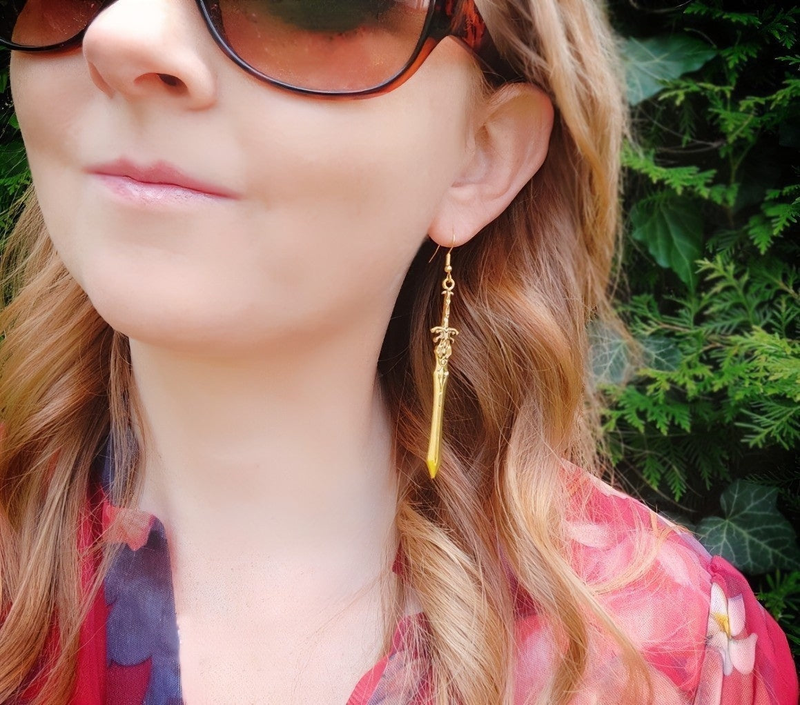 A female model wearing long slender gold sword earrings. They have a warm tone and look like rustic medieval weapons. 
