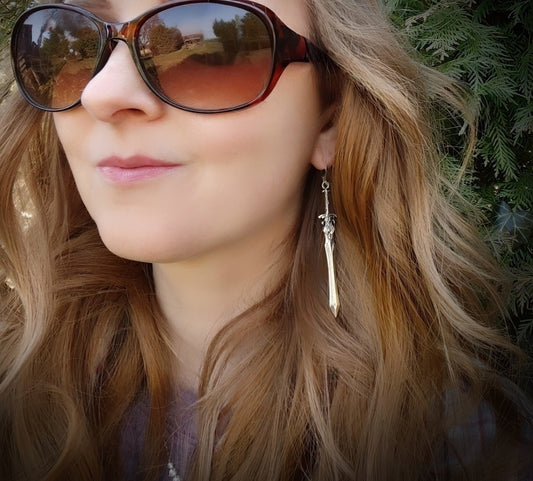 A female model wearing long silver sword earrings. She is outside in nature and the swords look cool dangling against her hair. 