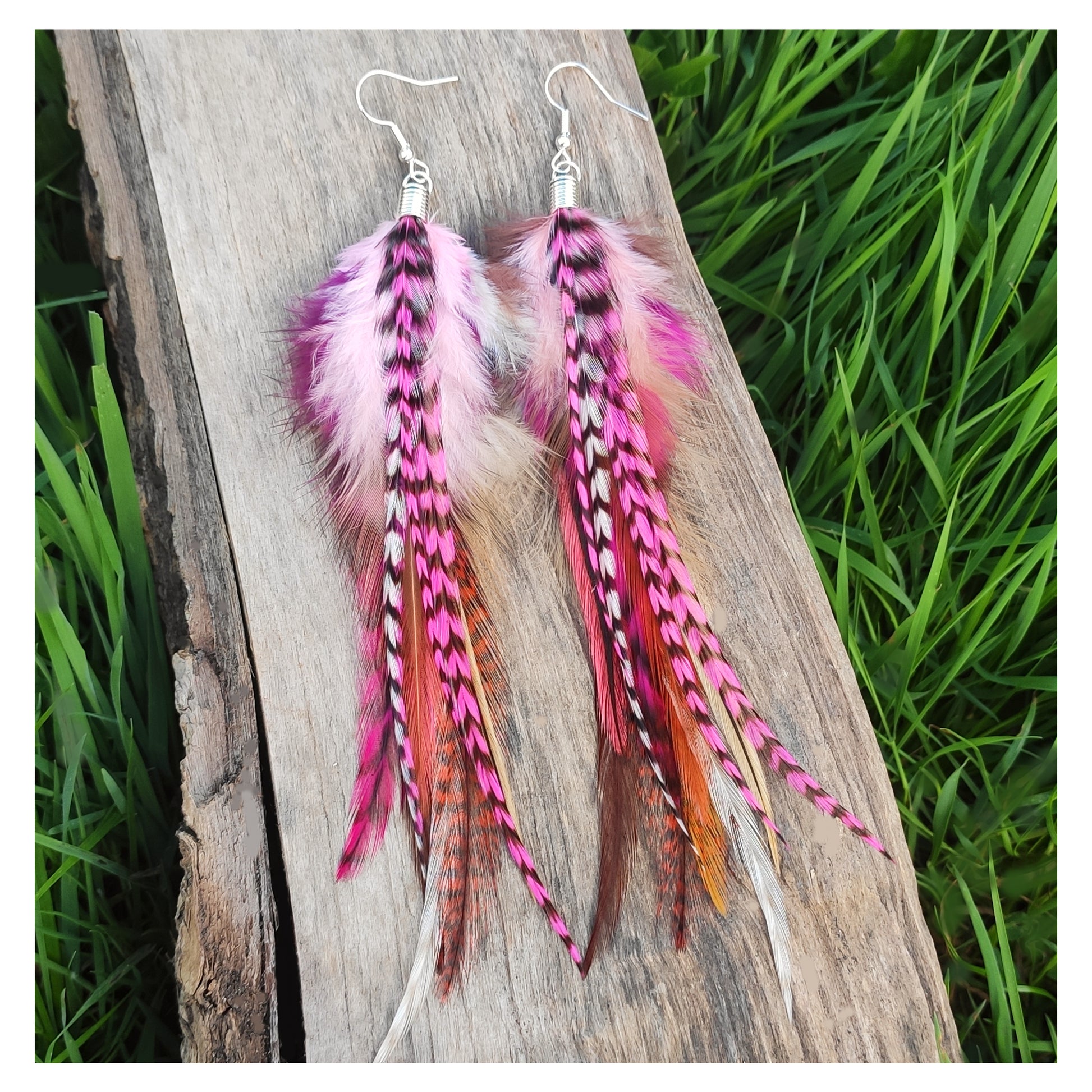Pink feather earrings on a wooden log with grass in the background. Stripy pink, white and brown feathers in a bohemian style.