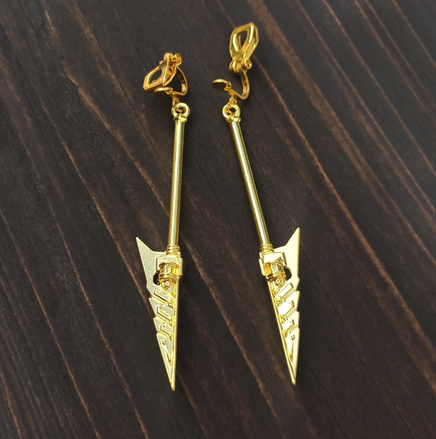 Gold arrow-shaped sword clip on earrings on a dark wooden surface. Fantasy mace earrings