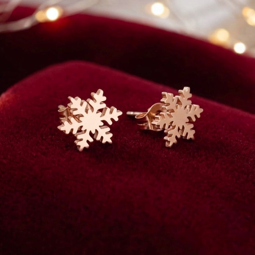 A pair of rose gold snowflake stud earrings resting on a deep red velvet jewelry box, with warm bokeh fairy lights blurred in the background.