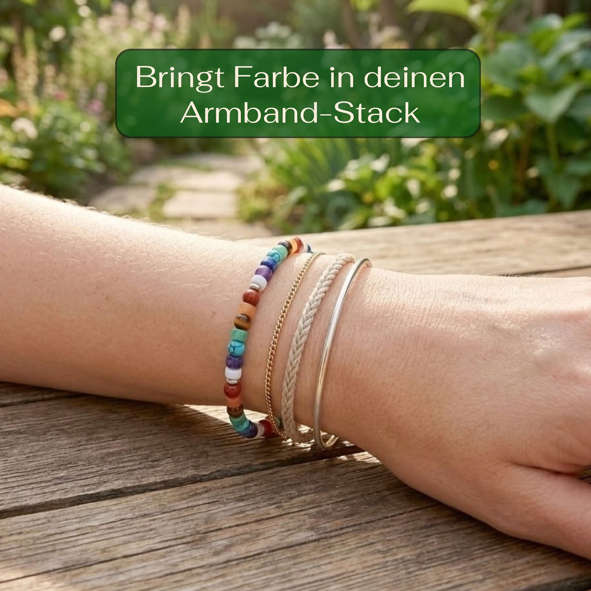 A close-up of a person's wrist wearing a stack of colorful beaded bracelets, a braided cord bracelet, and a thin silver bangle, resting on a wooden table with a blurred garden background. A green text box at the top reads 'Bringt Farbe in deinen Armband-Stack'.