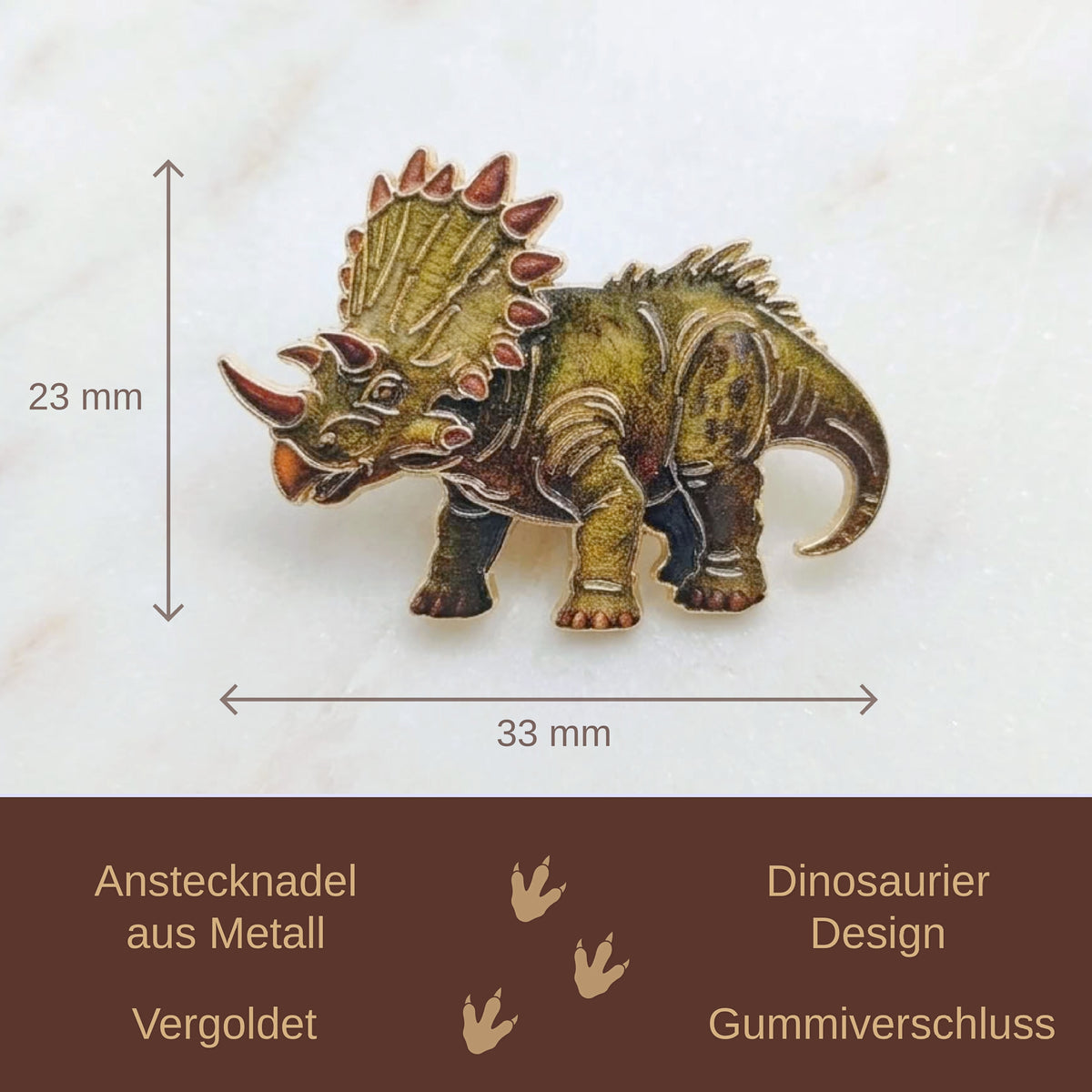 A small dinosaur-shaped metal pin or charm shown on a light marble-like background, with measurement arrows labeling it 23 mm tall and 33 mm wide. A dark brown banner at the bottom contains German text describing the product and features.