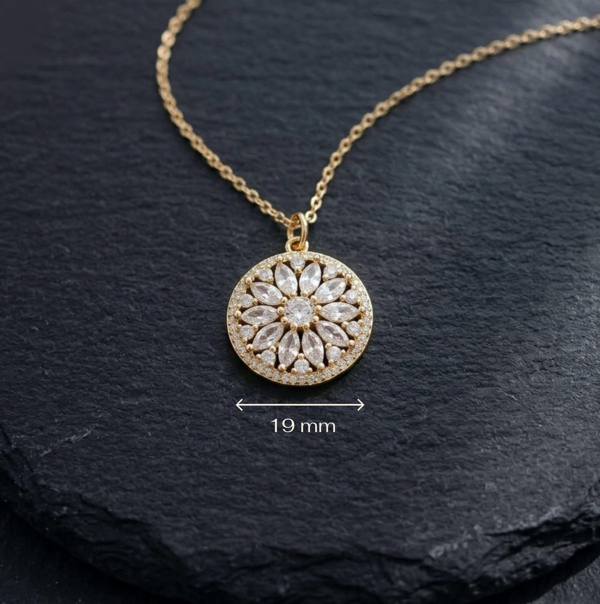 A gold circular pendant necklace with a daisy, sunflower or sunburst pattern of clear stones, displayed on a dark slate background with a '19 mm' size indicator overlay.