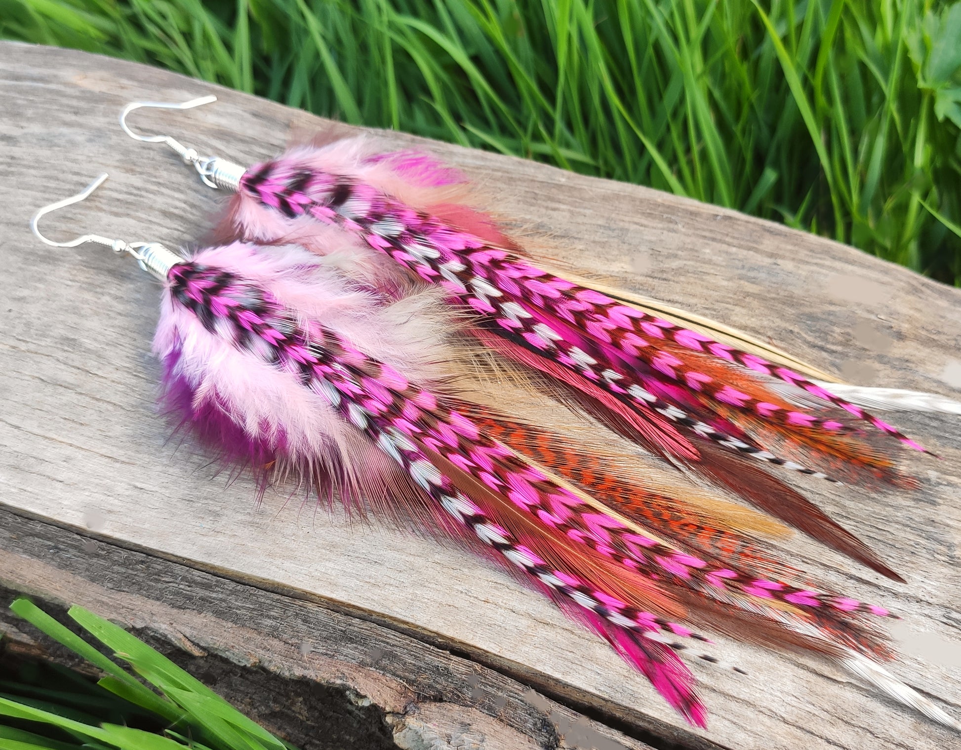 Feather earrings on a wooden surface with grass in the background. The feathers are long stripy pink white and brown.