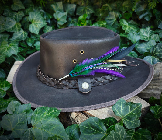 Brown leather cowboy hat with a colourful feather pin on a wooden surface with green leaves in the background