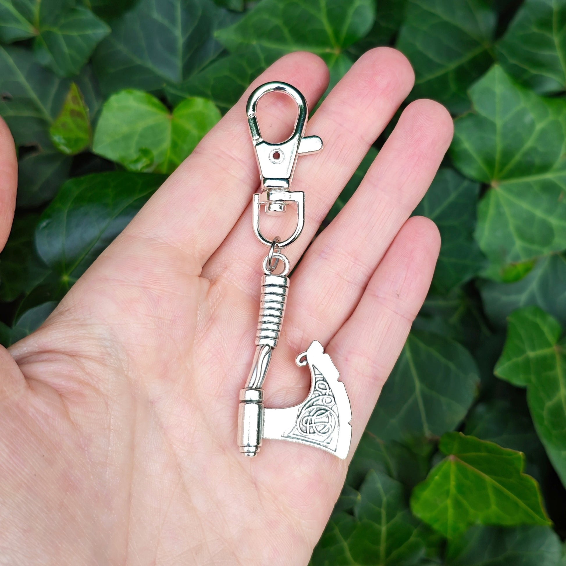 Hand holding a silver keychain with a axe and lobster clasp against a green leafy background