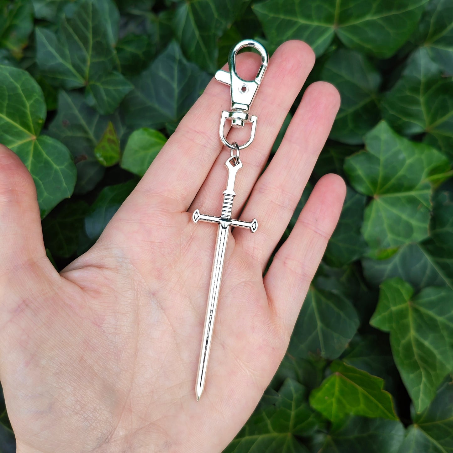 Silver sword keychain with lobster clasp held in a hand with a green leafy background