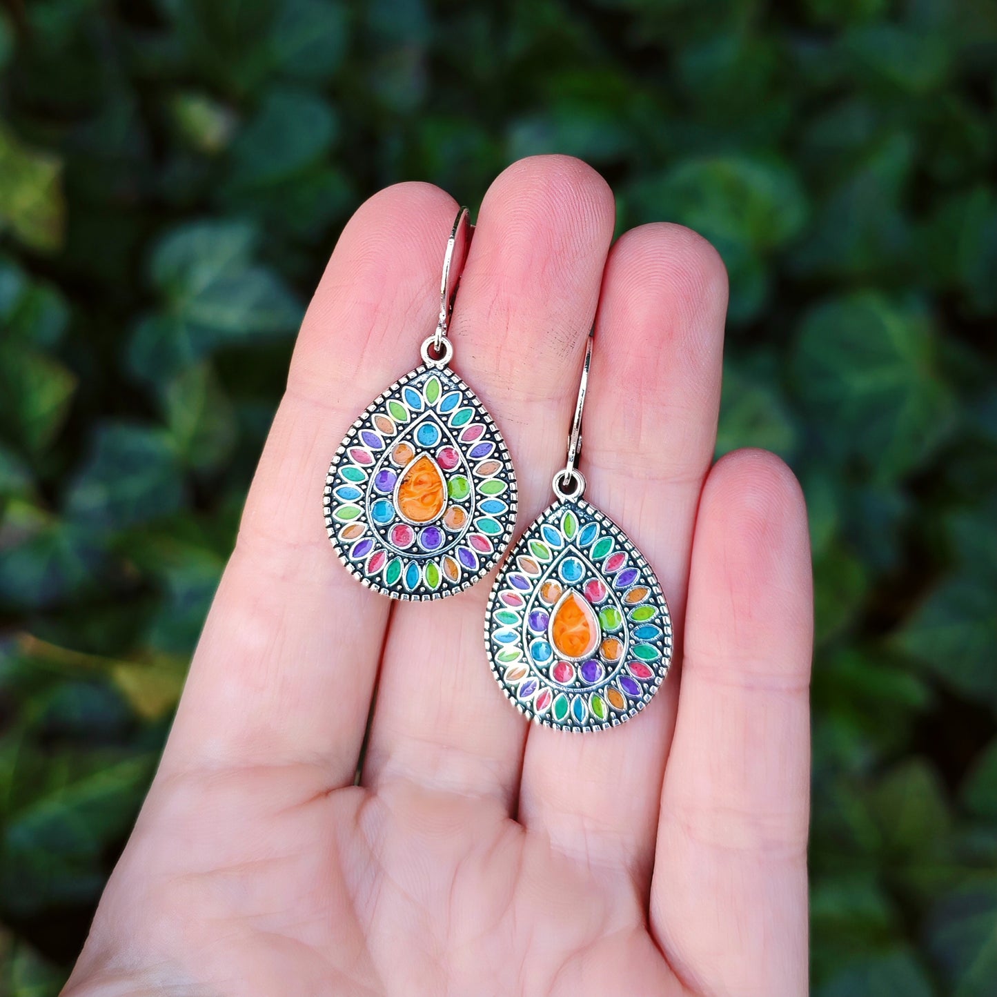 A hand holding colorful bohemian dangle earrings with silver hoops, and green leaves in the background