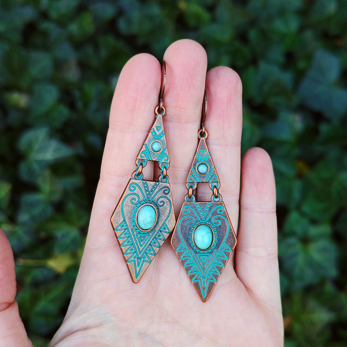 Turquoise and copper bohemian dangle earrings with intricate designs held in a hand against a green leafy background