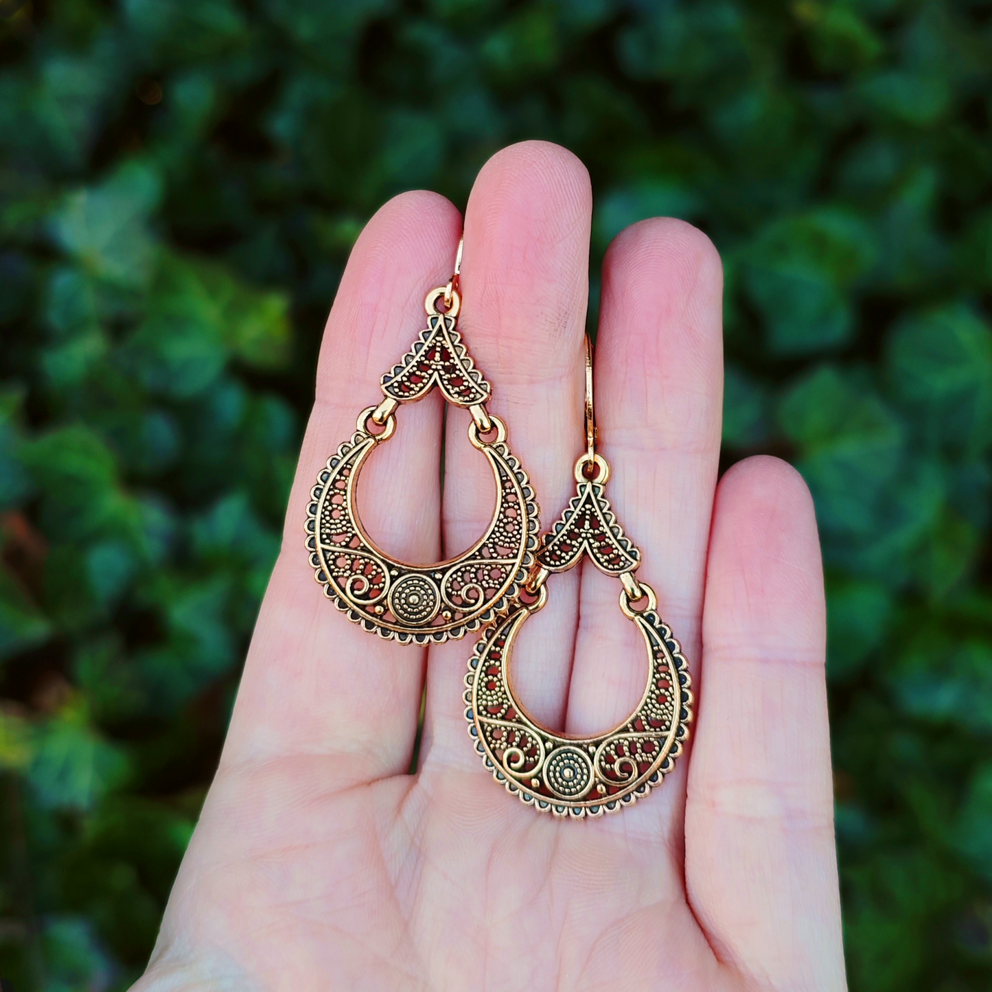 Gold filigree earrings held in a hand with a green leafy background