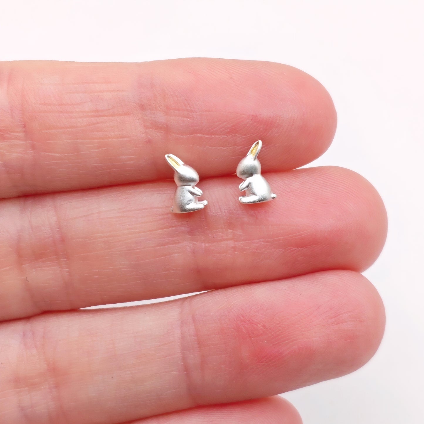 Small silver rabbit-shaped earrings with gold ears held between fingers on a white background