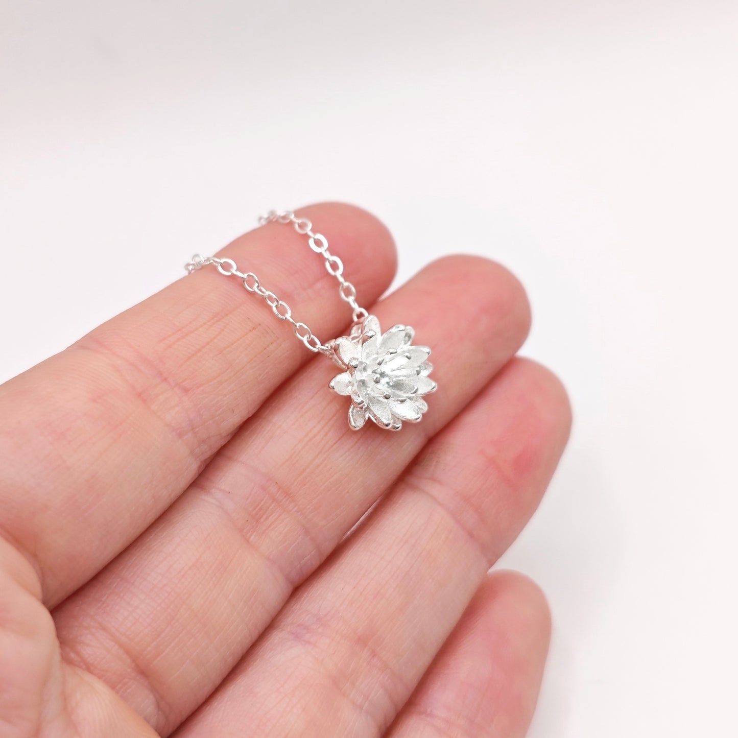 A hand holding a silver lotus flower necklace on a white bcakground