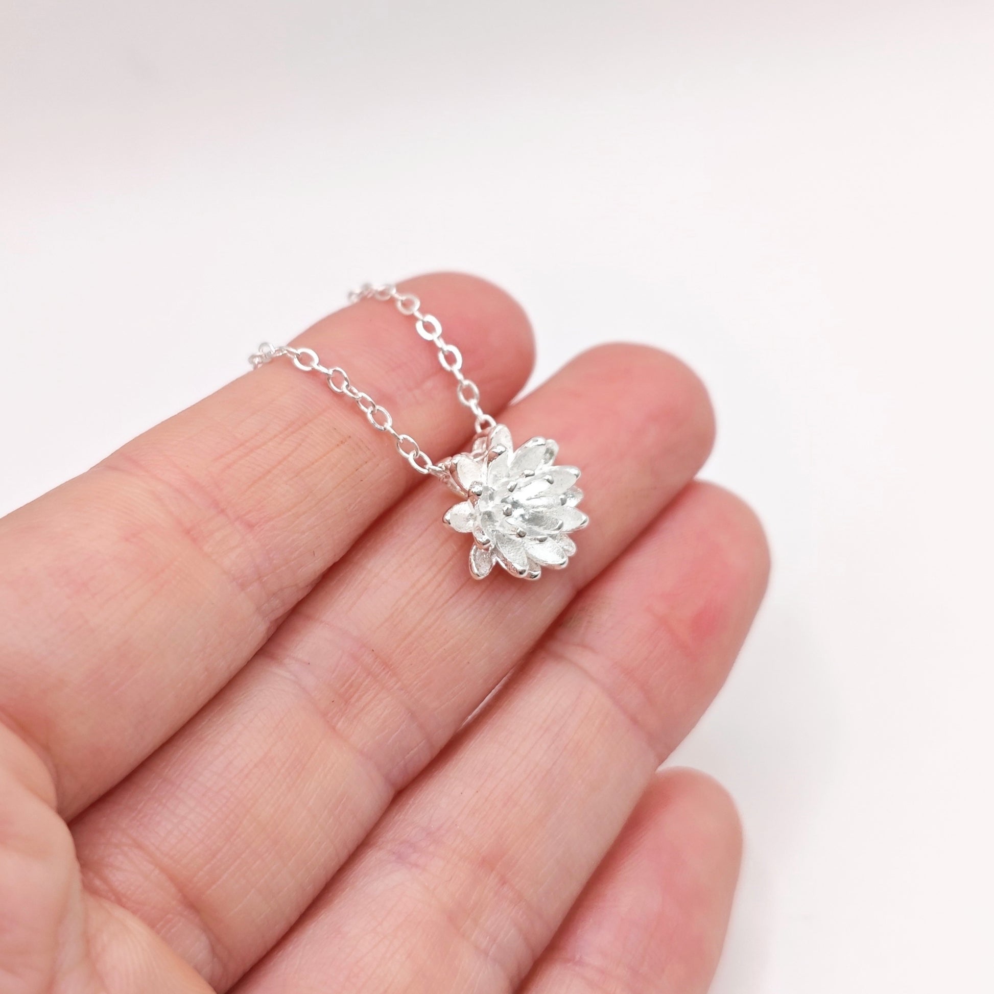 A hand holding a silver lotus flower necklace on a white bcakground