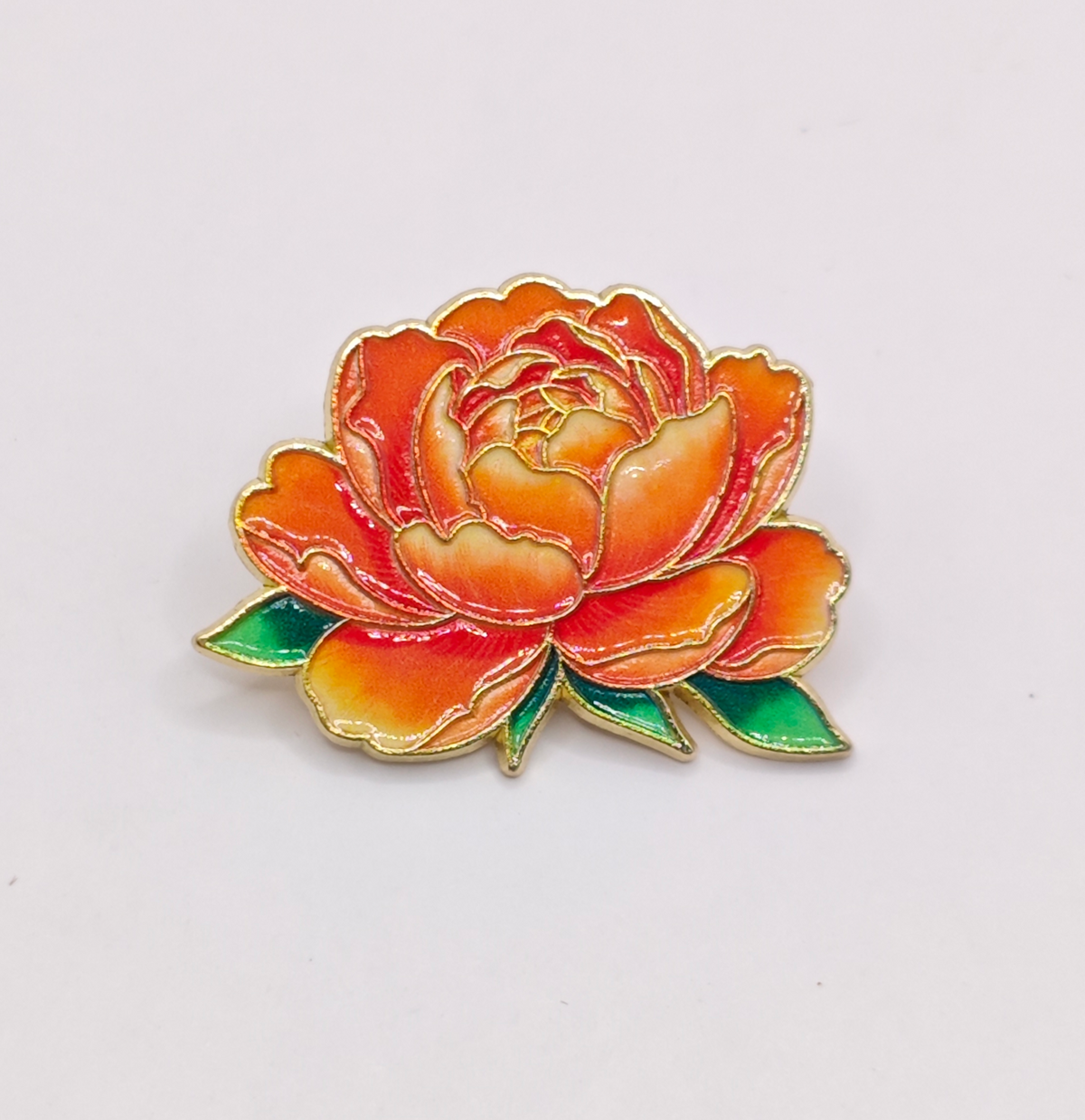 An enamel flower pin depicting an orange peony with green leaves, outlined in gold metal, on a plain white background.