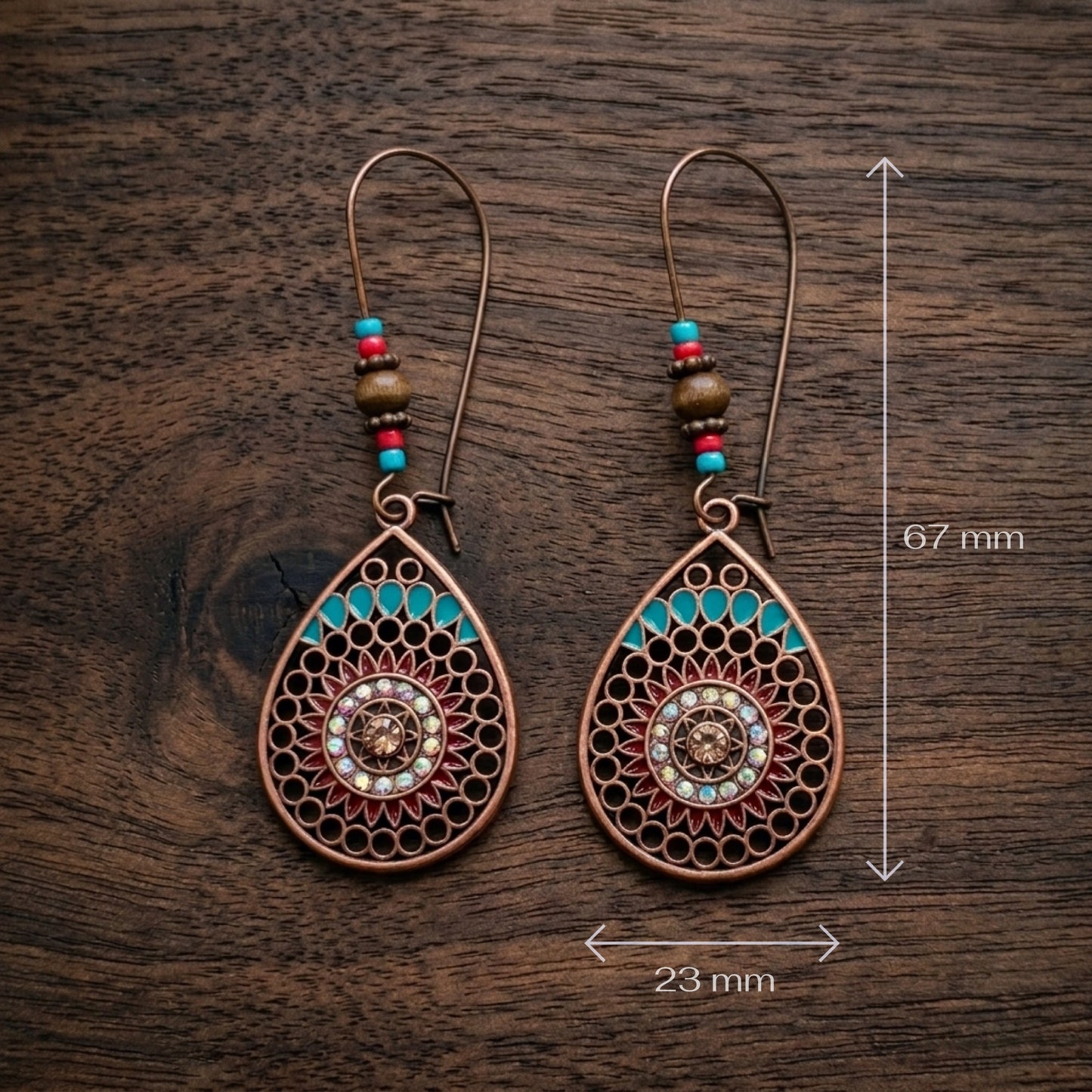 A pair of copper-toned teardrop dangle earrings with turquoise, red, and brown beads, shown on a dark wood background with dimension markers indicating 67 mm height and 23 mm width.