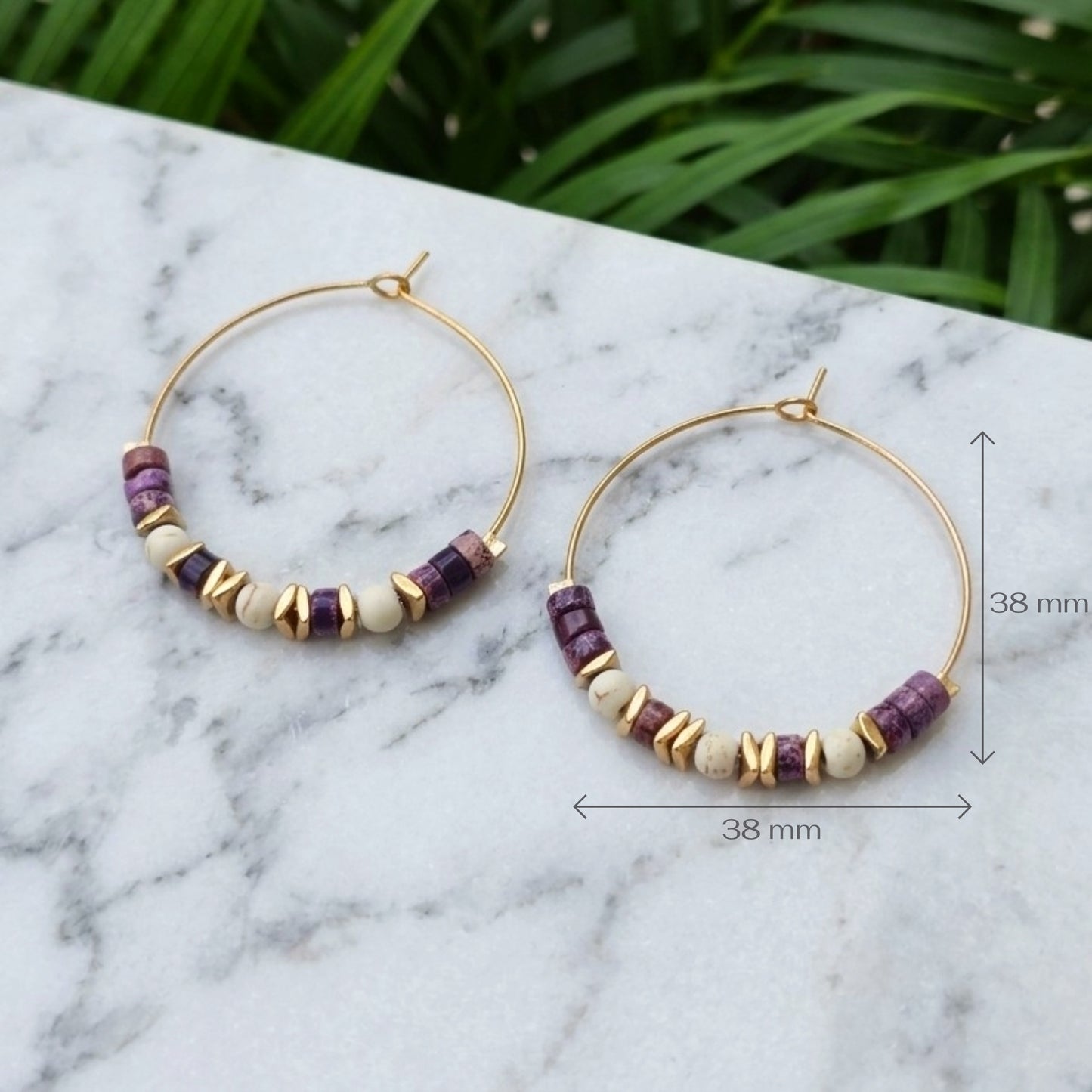 Gold hoop earrings with purple and cream beads, displayed on a white marble surface with green foliage in the background.