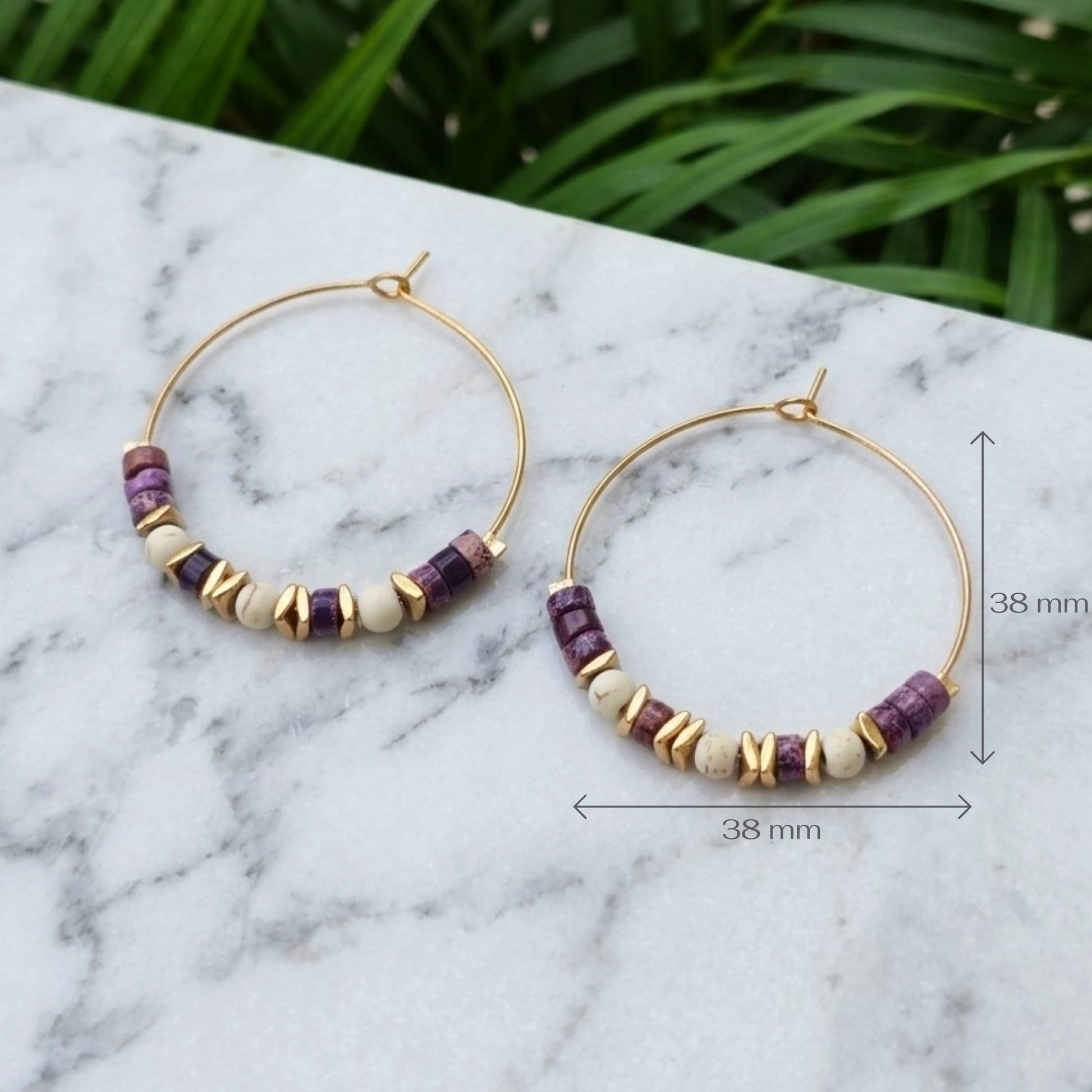 Gold hoop earrings with purple and cream beads, displayed on a white marble surface with green foliage in the background.