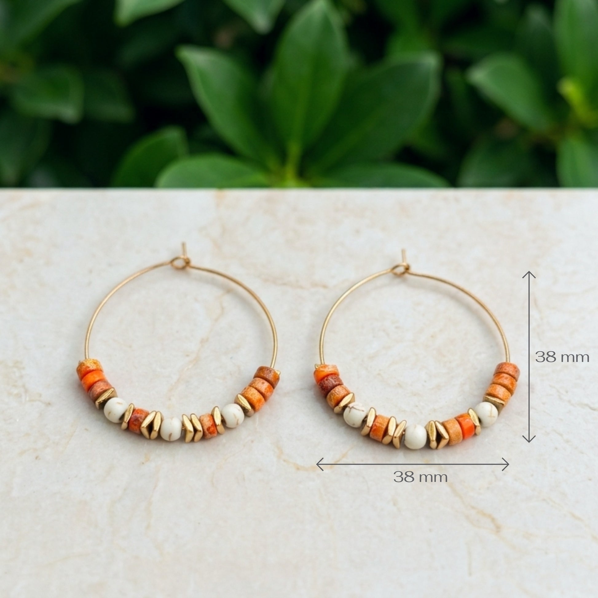 A pair of gold-tone hoop earrings with small orange, brown, and cream beads, displayed on a light marble surface with a blurred green plant background. Black dimension lines and text indicate a size of 38 mm.