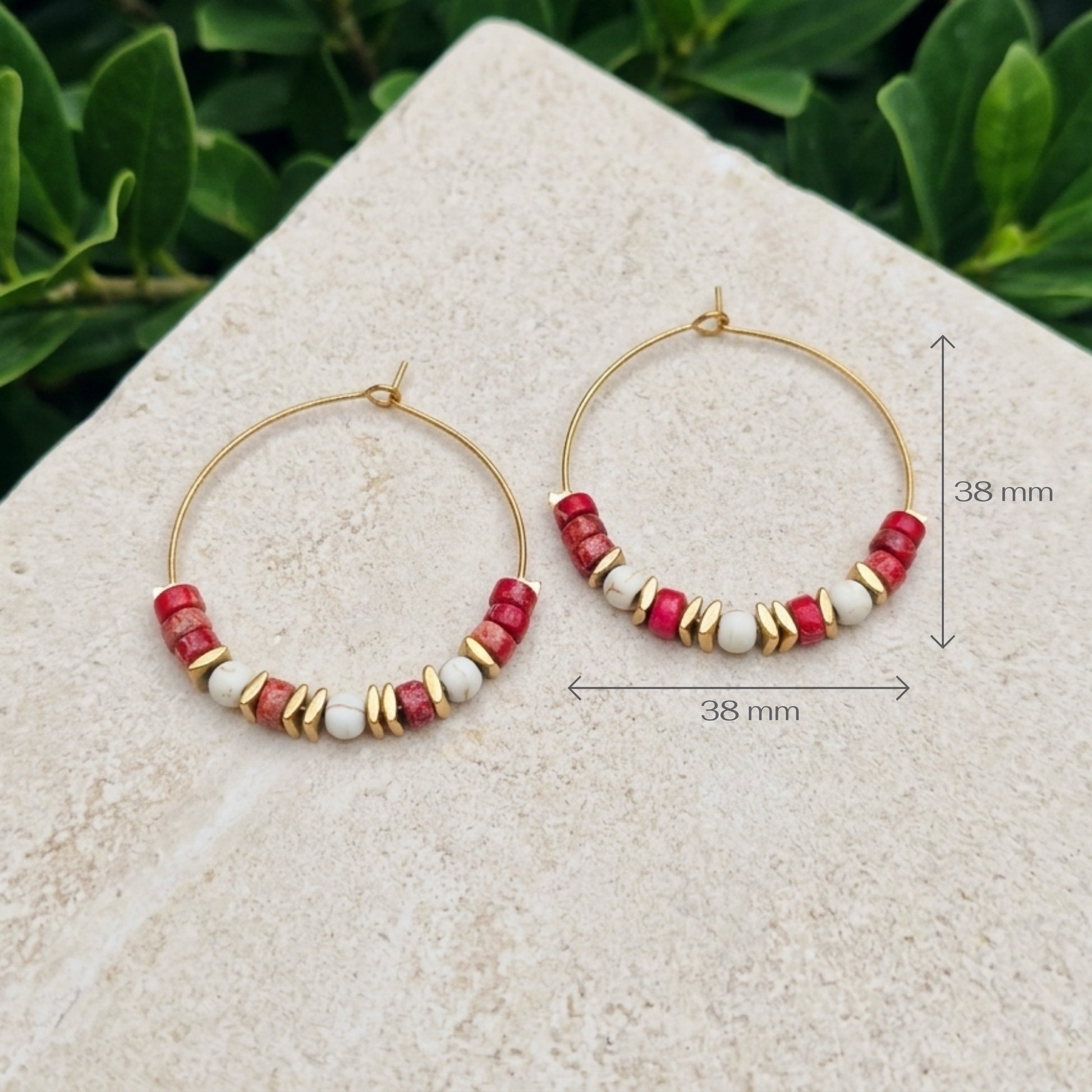 Gold hoop earrings with red sea jasper and white beads, displayed on a light stone surface with green foliage in the background. Measurement arrows and text indicate a size of 38 mm.