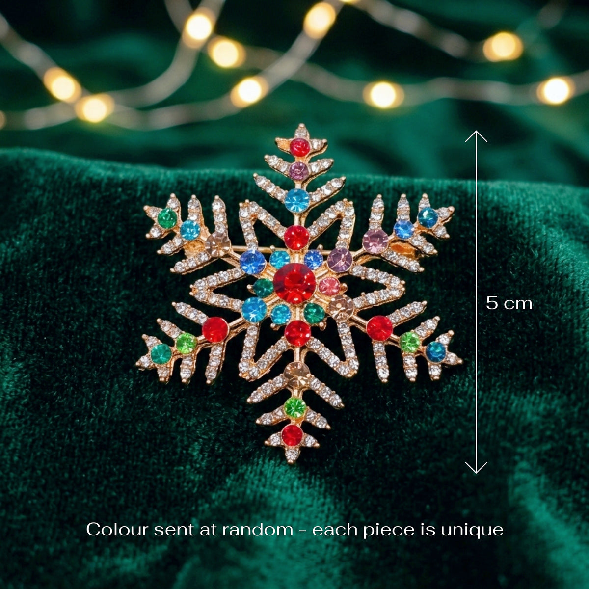 A gold snowflake-shaped brooch with multicolored Christmas gemstones, displayed on a dark green velvet background with blurred holiday lights. A vertical measurement arrow indicates the brooch is 5 cm tall, with text at the bottom stating 'Colour sent at random - each piece is unique'.