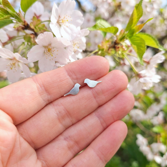 A hand holding brushed silver bird stud earrings with cherry blossom behind
