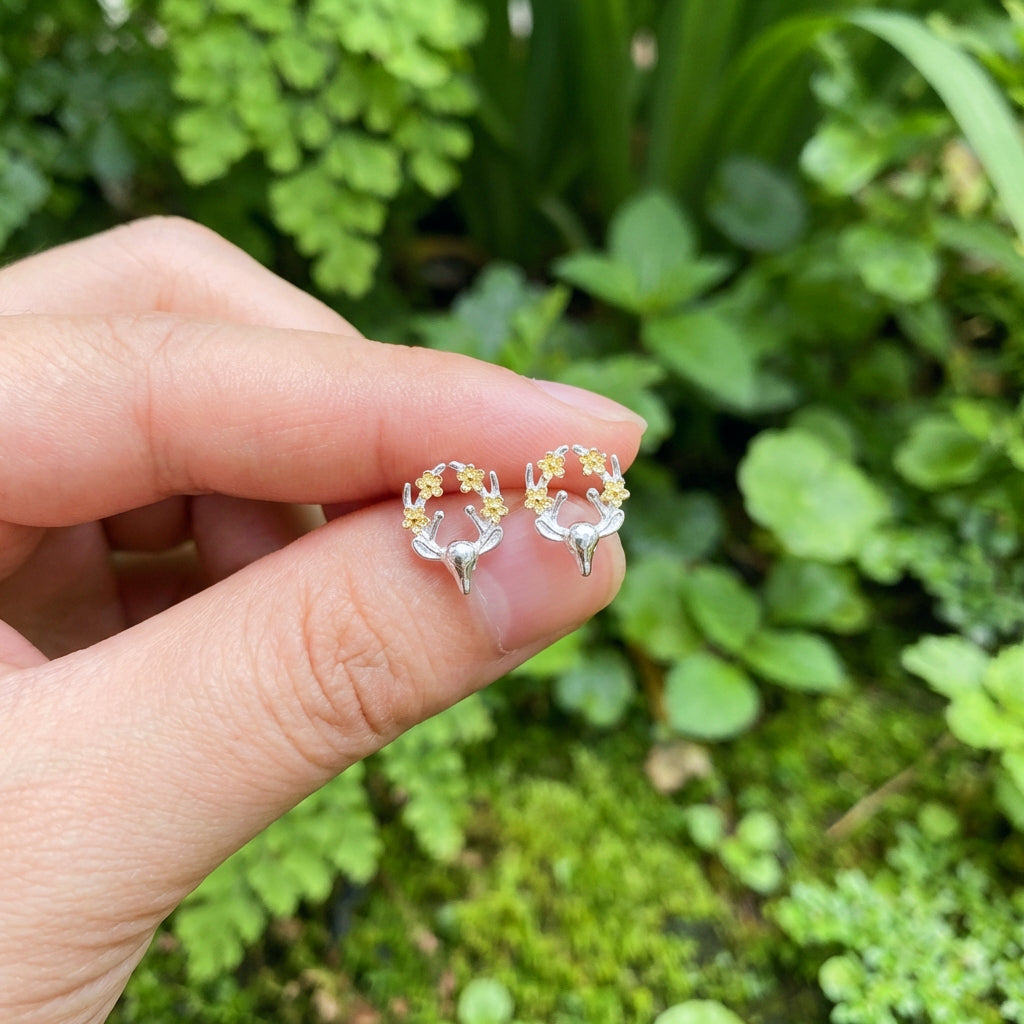 A hand holding silver and gold deer earrings with flowers on the antlers outside