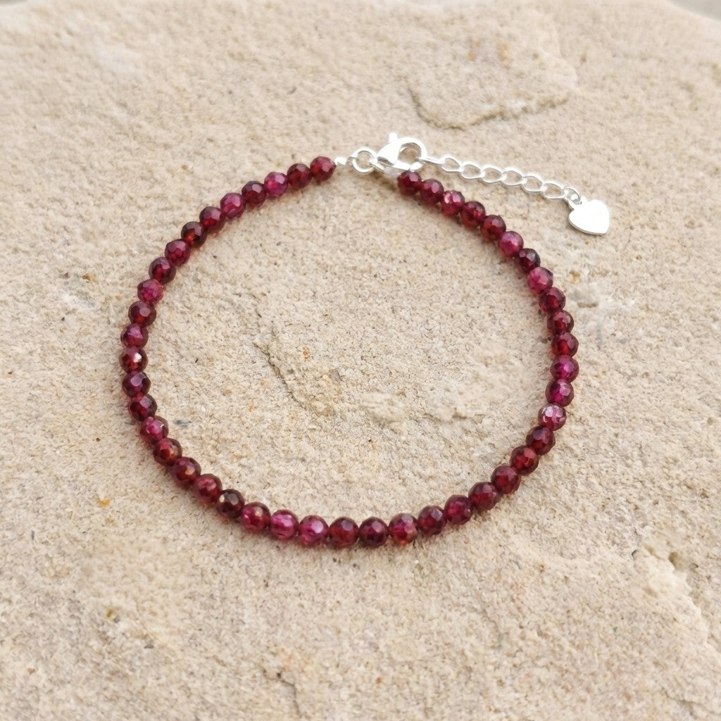 Dark red natural garnet gemstone bead bracelet on a light stone background. The beads are faceted and catch the light and the bracelet has an adjustable real 925 silver clasp.