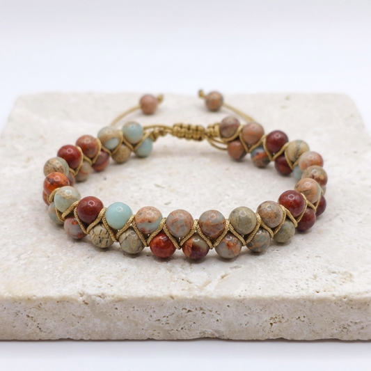 Sea sediment Jasper bead bracelet with a double layer of beads, woven together and an adjustable bolo drawstring. the beads are blue, red and beige with patterns. The natural gemstone bracelet is on a light stone background with greenery.