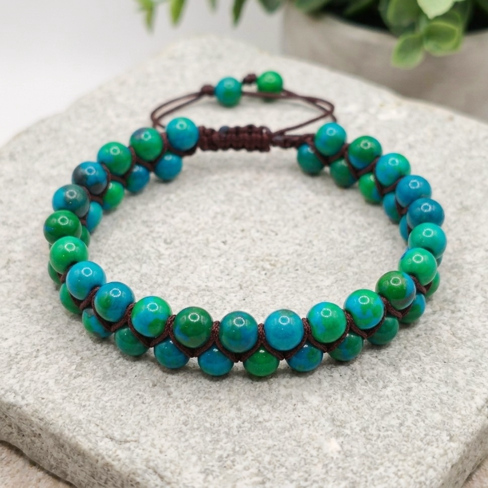 Chrysocolla bead bracelet featuring a double row of green and blue beads woven together with a brown cord and adjustable bolo drawstring. The natural colourful gemstone bracelet sits on a light stone background with greenery