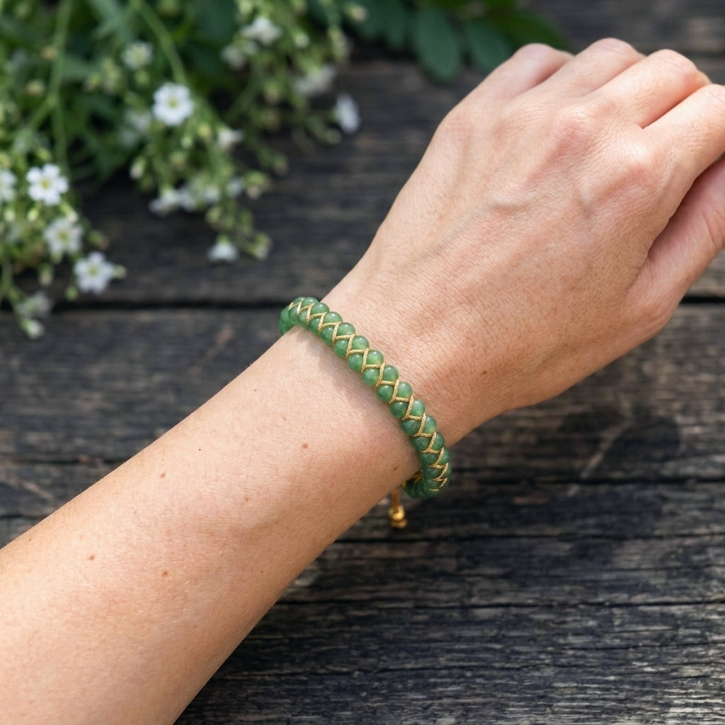 A woman's hand wearing a green aventurine braided bracelet on a wooden surface with flowers in the background