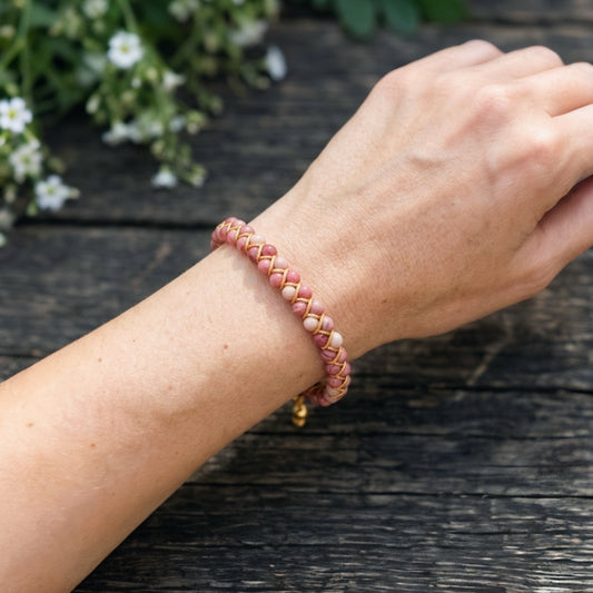 Hand wearing a pink stone braided bracelet on a wooden surface with white flowers in the background