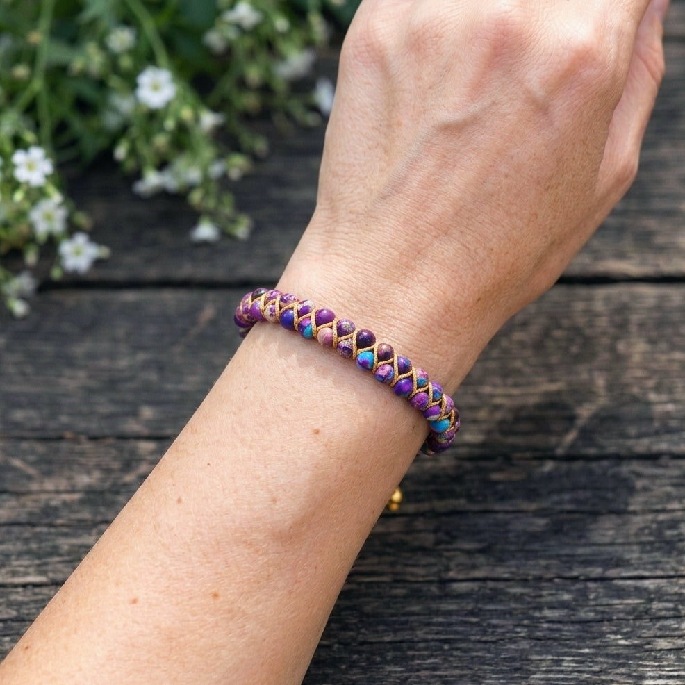 A woman's arm wearing a galaxy sea sediment jasper bead bracelet against a wood background. The colorful purple and blue gemstone beads are woven into a double row with an adjustable drawstring clasp.