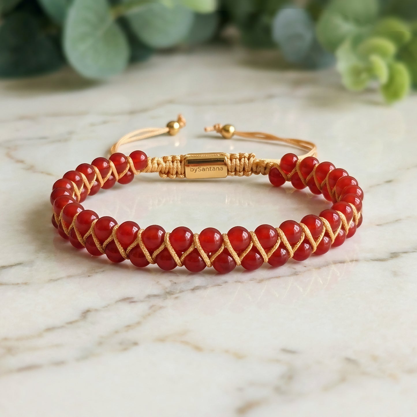Bead bracelet with red agate gemstone beads in a double row, woven with an adjustable drawstring cord, on a marble surface with greenery in the background