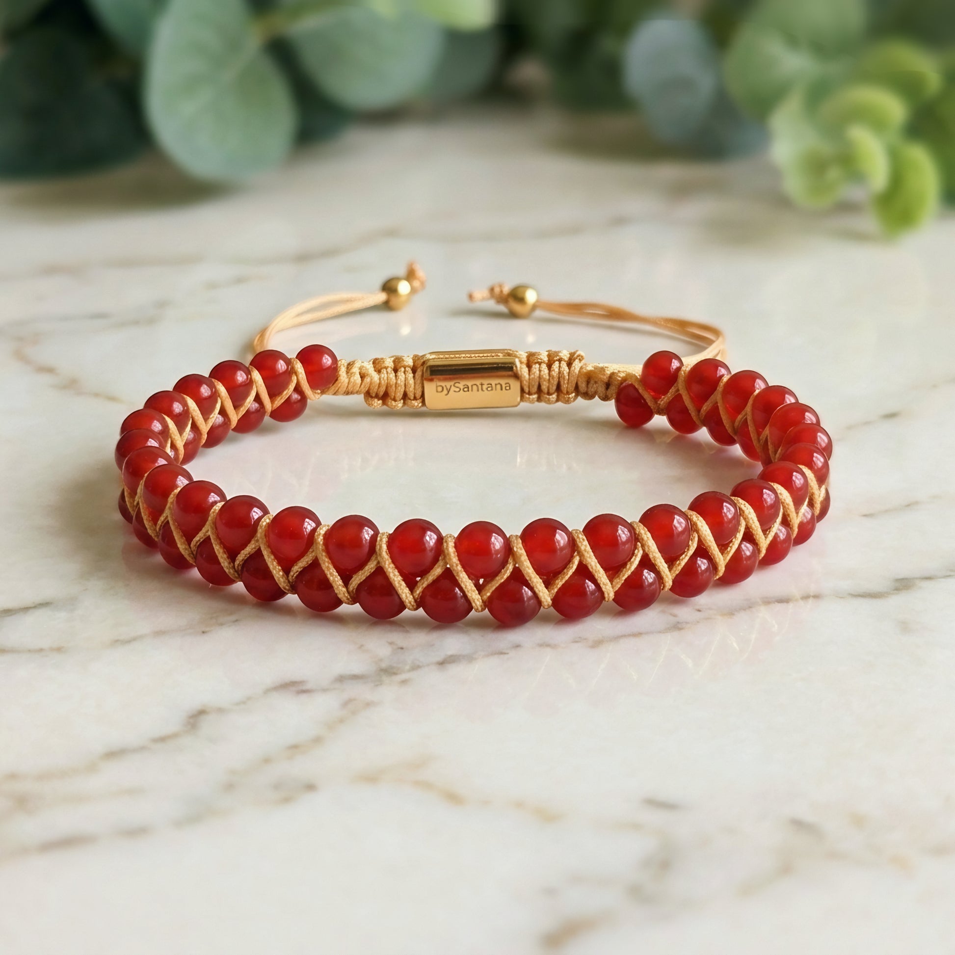 Bead bracelet with red agate gemstone beads in a double row, woven with an adjustable drawstring cord, on a marble surface with greenery in the background