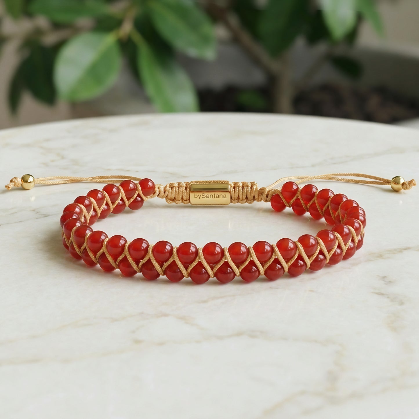 Bead bracelet with red agate gemstone beads in a double row, woven with an adjustable drawstring cord, on a marble surface with greenery in the background