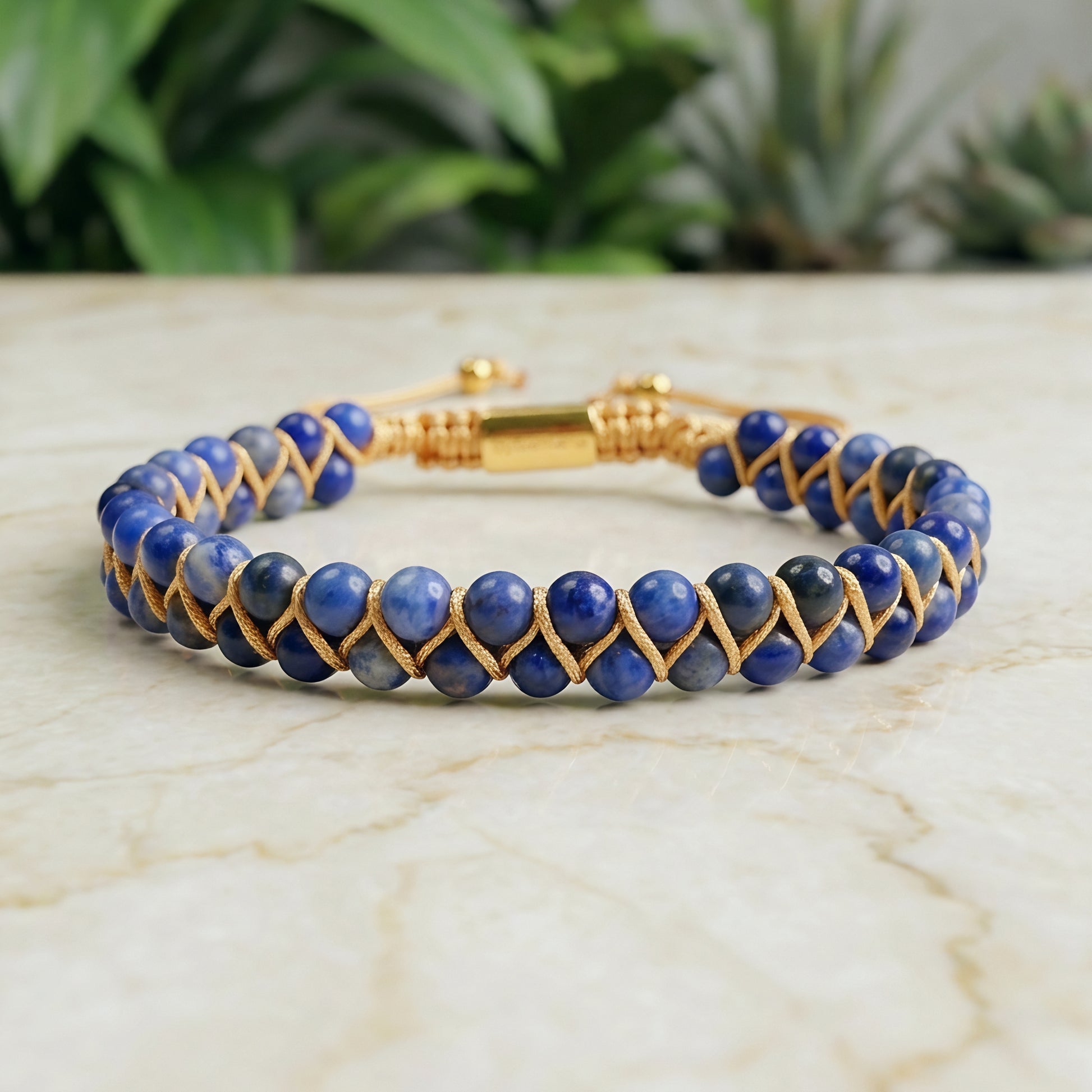 Blue sodalite beaded gemstone bracelet with gold cord on a marble surface with greenery in the background