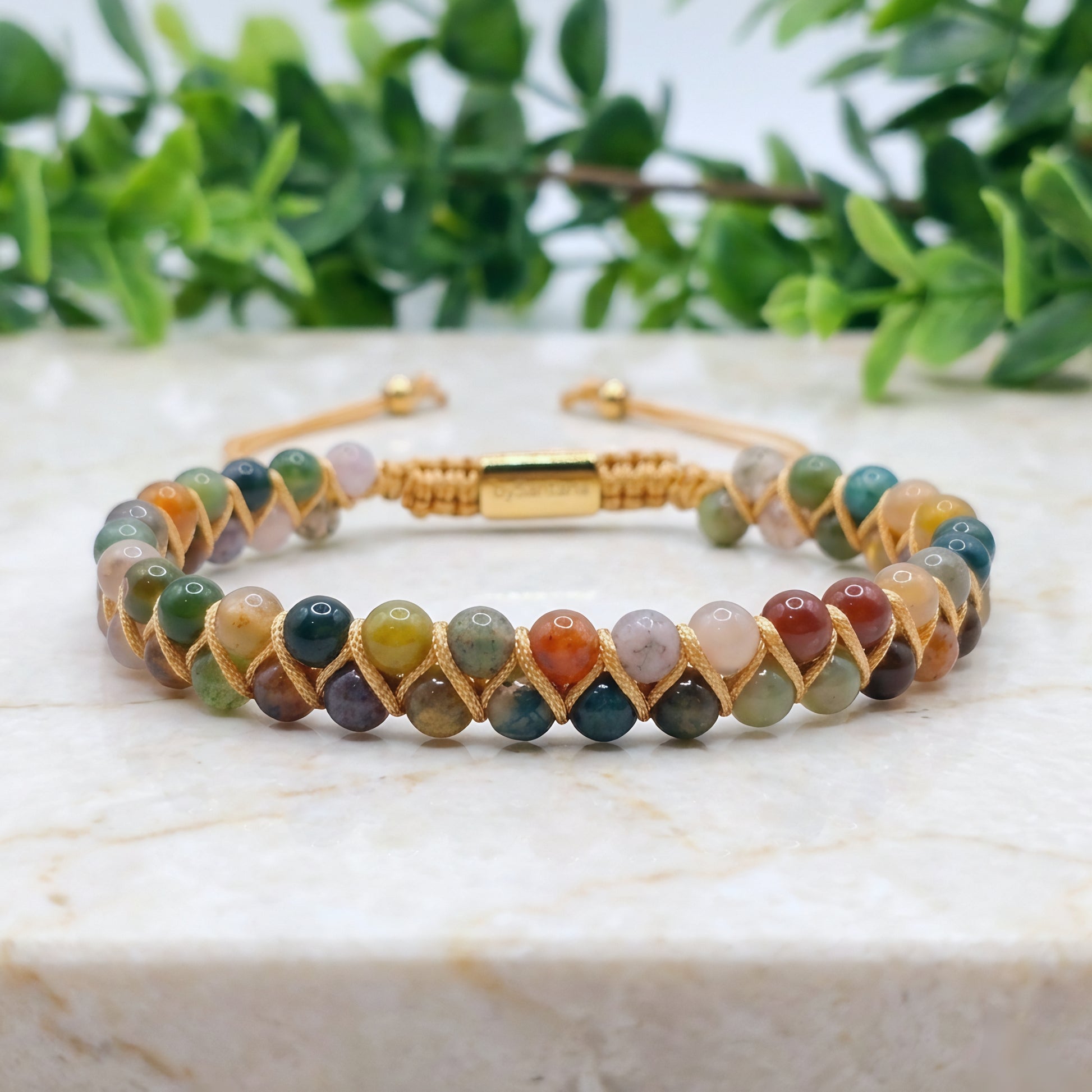 Multicolored beaded bracelet with Indian Agate gemstone beads and gold cord wrapping on a marble surface with greenery in the background