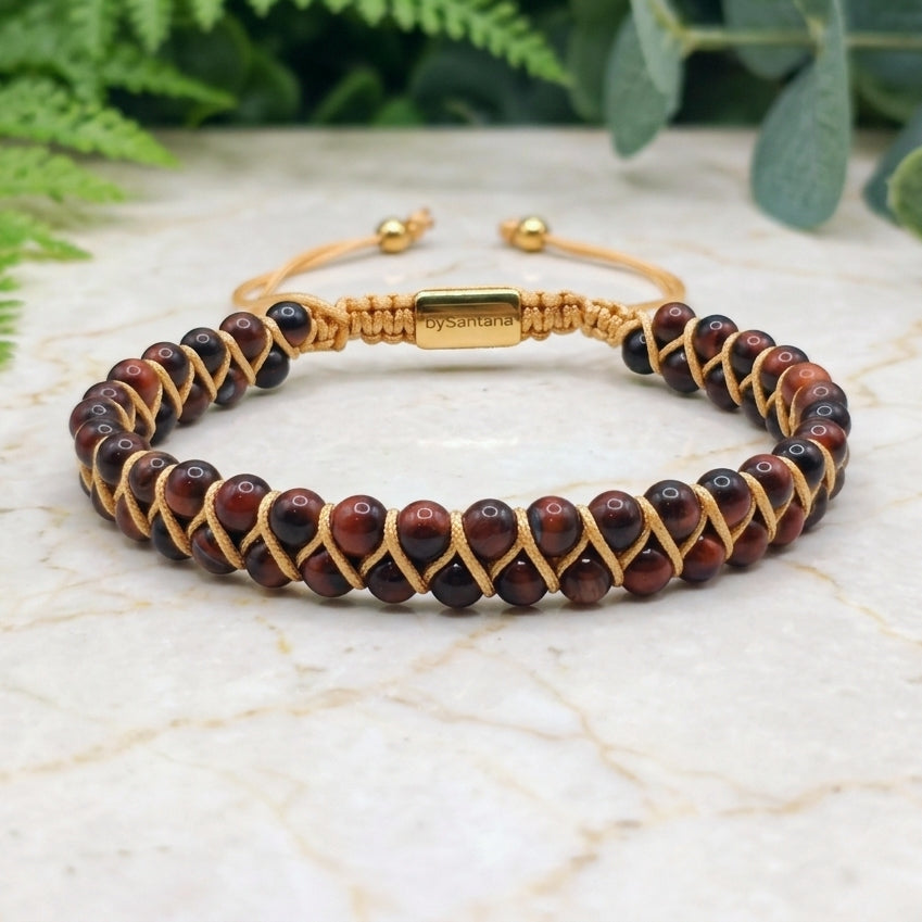 Bracelet with red tiger's eye gemstone beads and gold braided cord on a marble surface with green leaves in the background