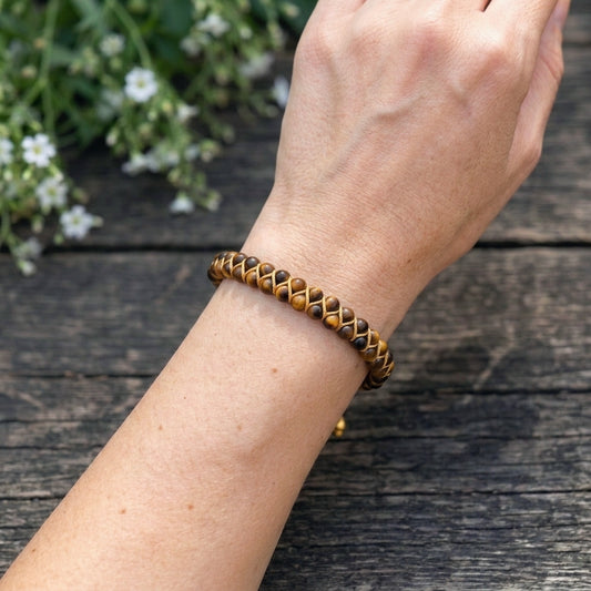 A woman's hand wearing a gold tigers eye beaded bracelet on a wooden surface with white flowers in the background