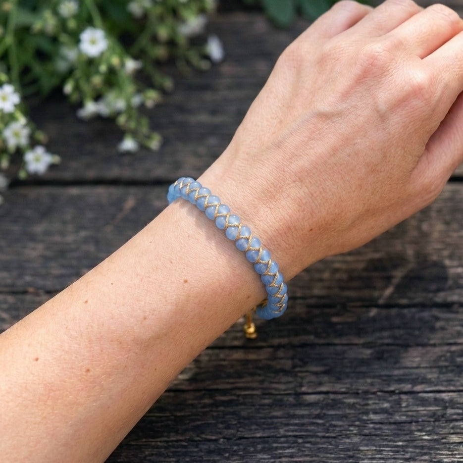 A woman's hand wearing a blue aquamarine braided bracelet on a wooden surface with white flowers in the background