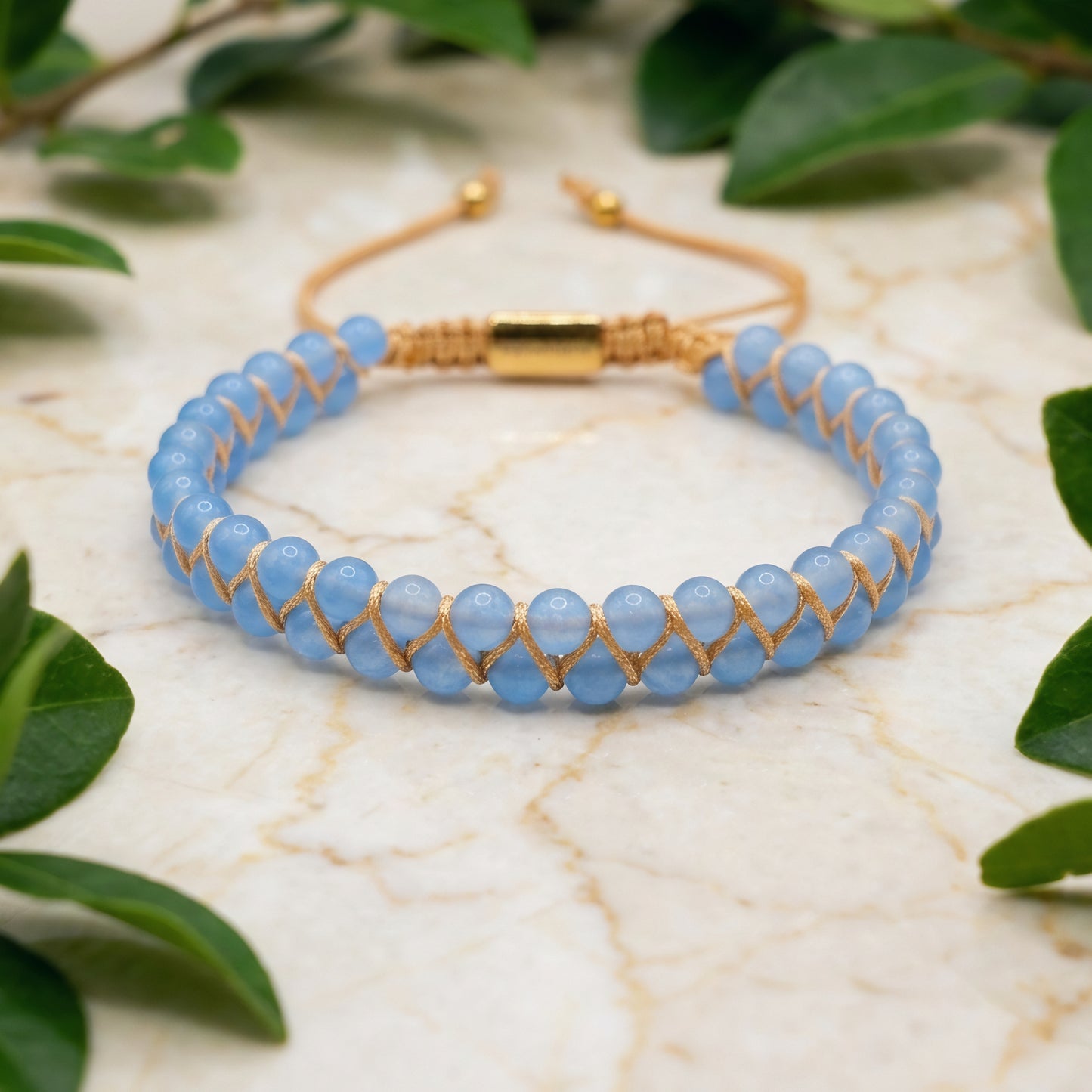 Blue aquamarine beaded gemstone bracelet with gold accents on a marble surface with green leaves.