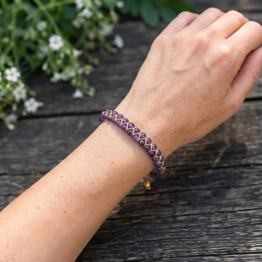 A woman's hand wearing a purple amethyst braided bracelet on a wooden surface with white flowers in the background