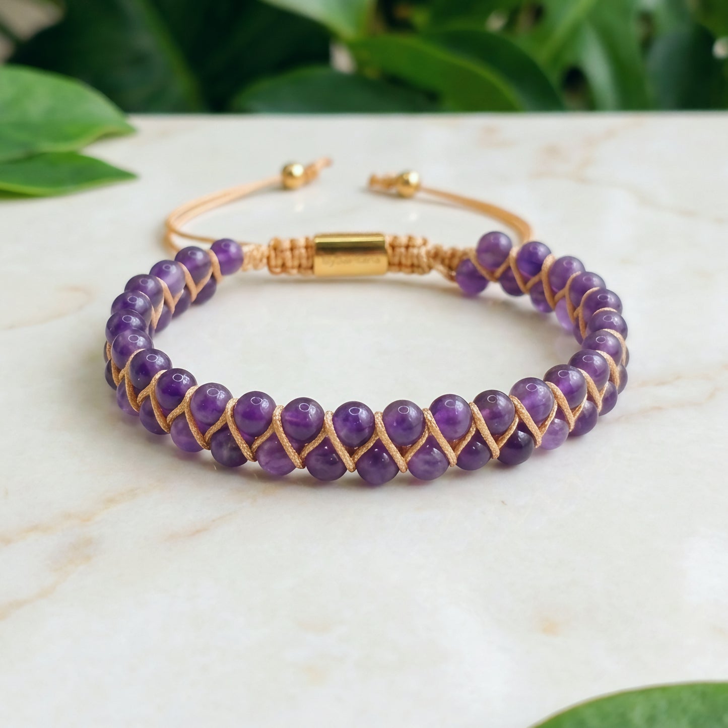 Purple beaded bracelet with gold accents on a marble surface with green leaves in the background