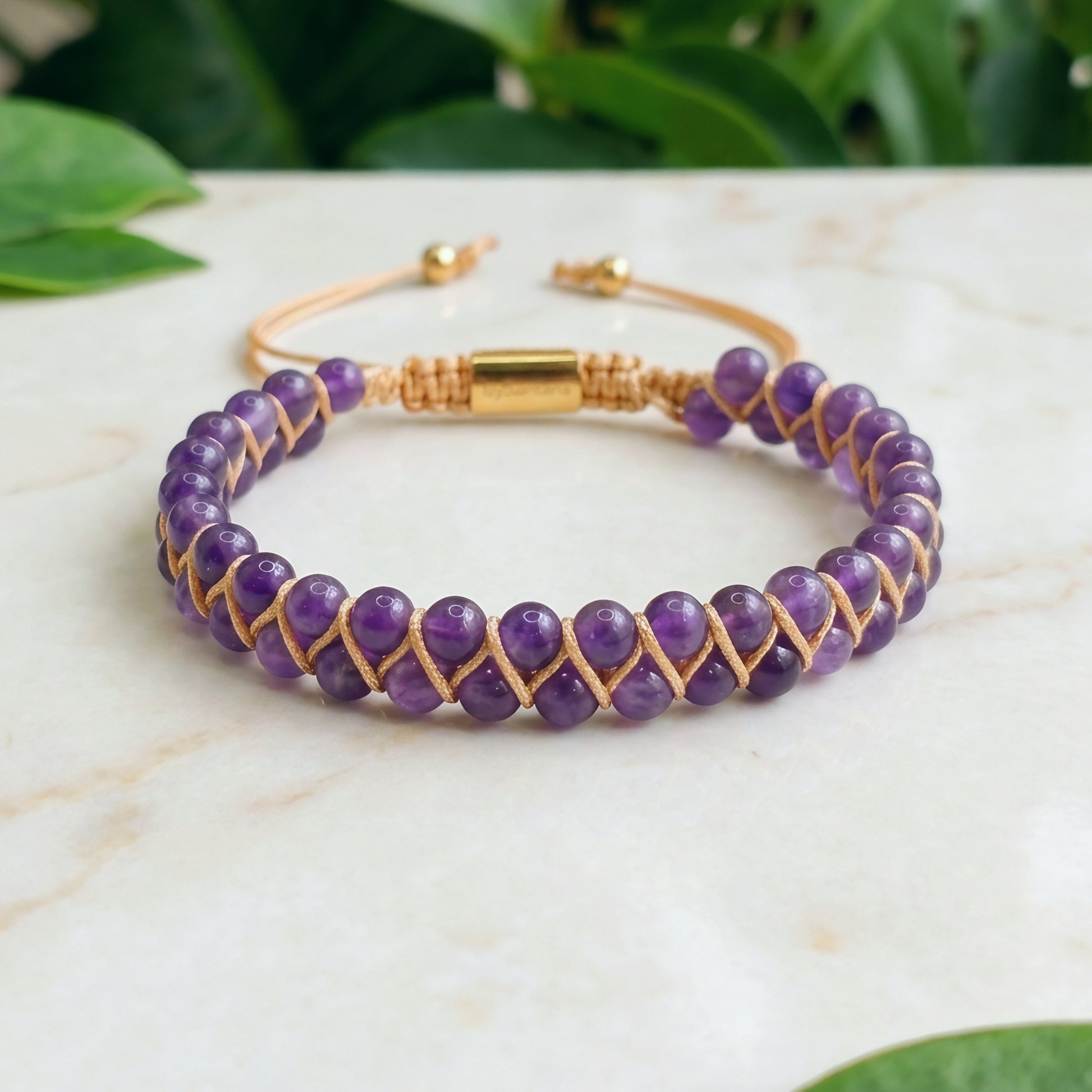 Purple beaded bracelet with gold accents on a marble surface with green leaves in the background