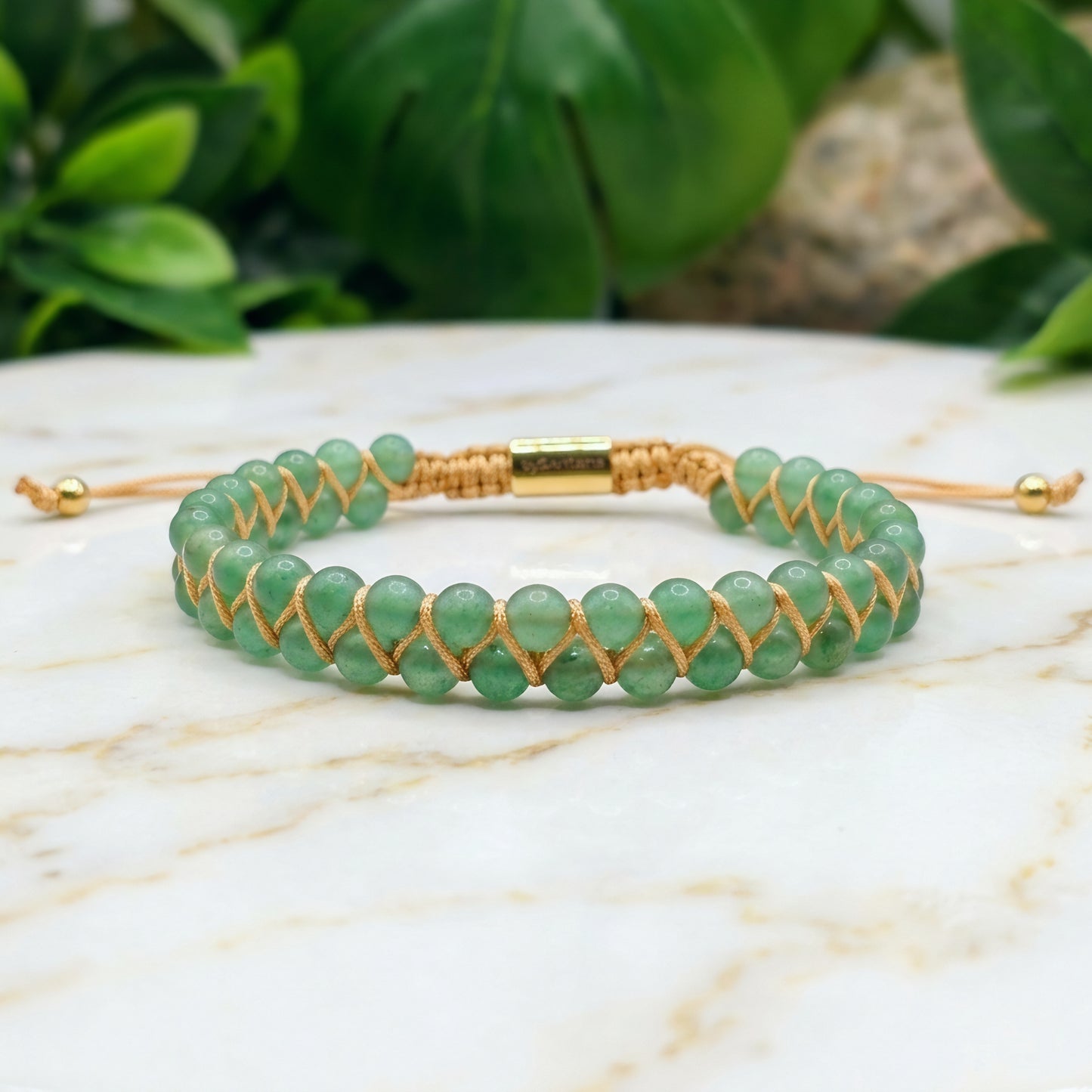 Green aventurine beaded gemstone bracelet with gold cord on a marble surface with green leaves in the background
