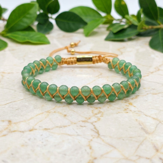 Green aventurine beaded gemstone bracelet with gold cord on a marble surface with green leaves in the background