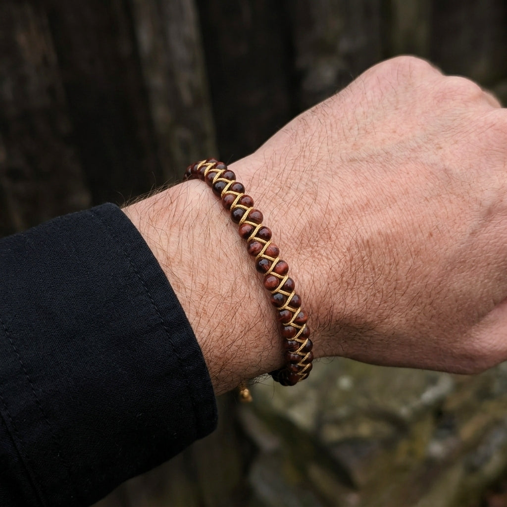 Close-up of a man's wrist wearing a braided red tigers eye bracelet with a blurred natural background.